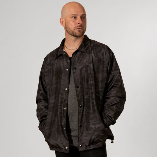 A black camouflage coaches jacket features a relaxed fit and a snap-button closure. It has a branding logo on it.
