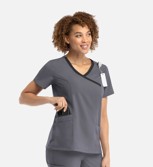 A woman in a grey mock wrap top with contrast trim, featuring a pocket. The top has a logo and a tag attached.