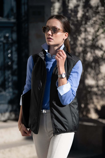 A woman wearing a black unisex vest over a blue shirt, styled with sunglasses and light-coloured trousers.