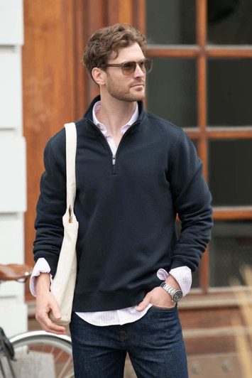 A navy unisex sweatshirt with a zip collar, worn by a man carrying a tote bag. The background features wooden structures.