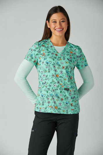 A floral print scrubs top in mint green, featuring a variety of colourful designs, worn over a light blue long-sleeve shirt.
