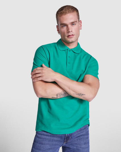 A green Austral Polo Shirt worn by a model with short hair, standing with arms crossed. The shirt has a collar and features a logo.