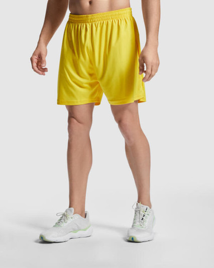 Yellow sports shorts with an elastic waistband, worn by a model alongside white athletic shoes.