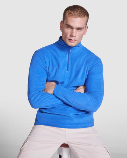 A blue polar fleece jumper with a half zip, featuring a logo, modelled by a seated male figure.