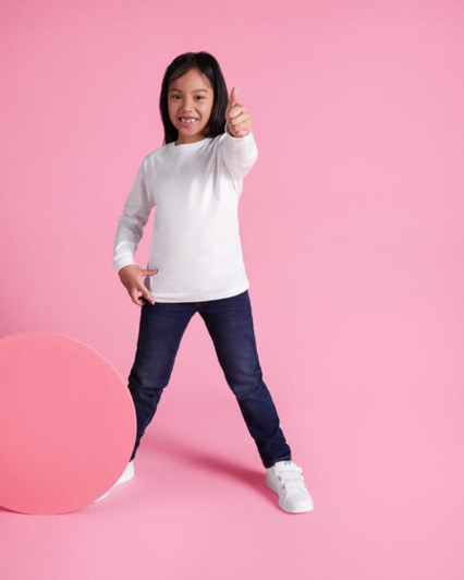 A child poses in a white long-sleeve t-shirt and blue jeans against a pink background, giving a thumbs-up.