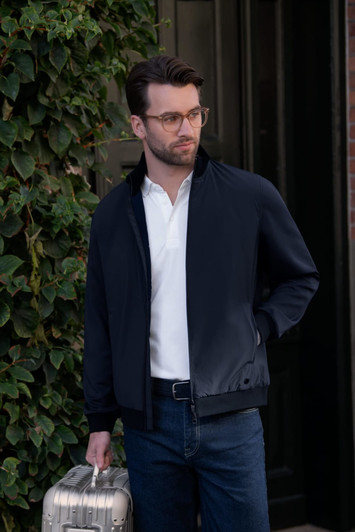 A man wears a navy hybrid jacket, holding a silver suitcase, standing by an ivy-covered wall. The jacket has a logo.