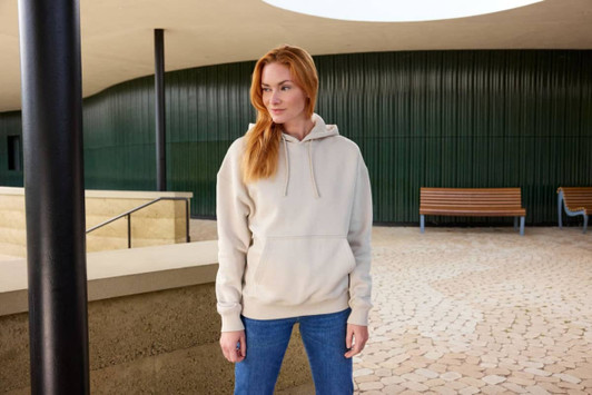 An oversized unisex sweatshirt in beige, featuring a hood, worn with blue jeans in an outdoor setting.