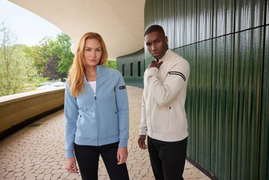 A light blue ladies zip jacket and a cream men's zip jacket with a logo, displayed against a modern architectural background.