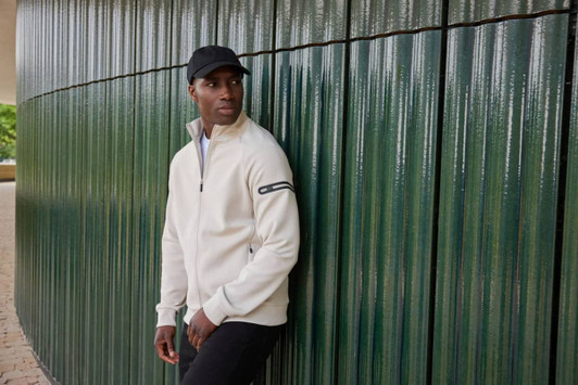 A men's zip jacket in cream, featuring a logo, worn by a model against a textured green wall.