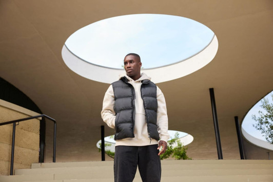 A man wears a black puffer vest over a beige hoodie, posing outdoors under an architectural structure.