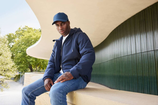A man sits outside wearing a navy bomber jacket and a matching cap, paired with jeans. The background features greenery and modern architecture.