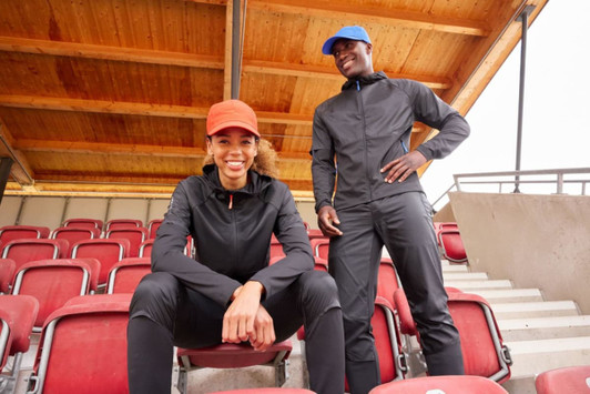 A black ladies running jacket is worn by a woman sitting, while a man in a matching black outfit stands beside her.