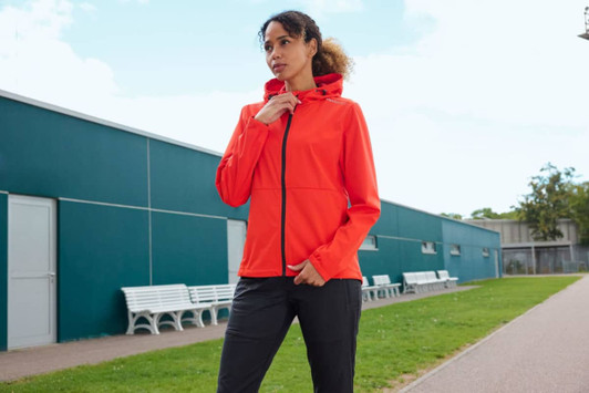 A bright orange softshell jacket with a hood, worn by a woman outdoors, near a green lawn and buildings.
