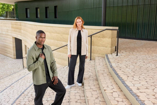 A unisex shirt jacket in olive green and beige worn by models on stone steps, featuring a logo.
