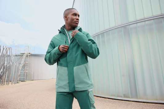 A model wearing a green hardshell rainjacket, featuring contrasting shades and a logo, set in an outdoor industrial environment.