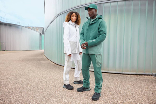 A green hardshell rainjacket is worn by a man, alongside a woman in a white rainjacket, set outdoors.