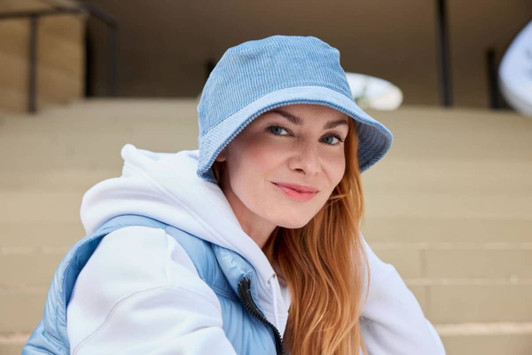 A corduroy bucket hat in light blue, worn by a woman with long hair, seated outside. It features a logo.