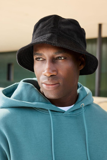 A black corduroy fisherman hat sits atop a model wearing a green hoodie, displayed against an outdoor background.