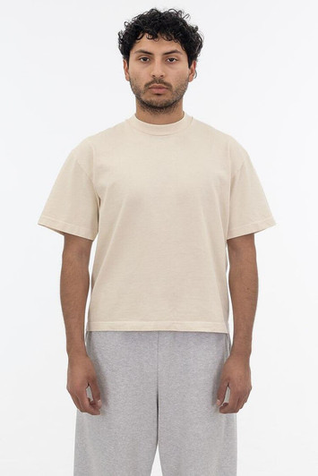 A short sleeve garment dye t-shirt in a beige colour, worn by a model with curly hair and grey pants.