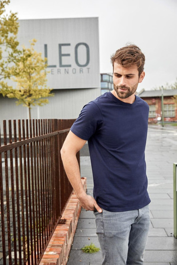 A navy blue T-shirt worn by a man standing outdoors near a fence and a building in a urban setting.