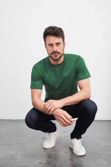 A green T-shirt worn by a man squatting in jeans and white sneakers, featuring a logo.