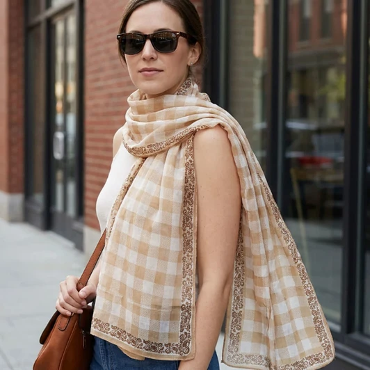 A woman wearing a lightweight beige and cream checkered scarf, paired with sunglasses and carrying a brown bag.