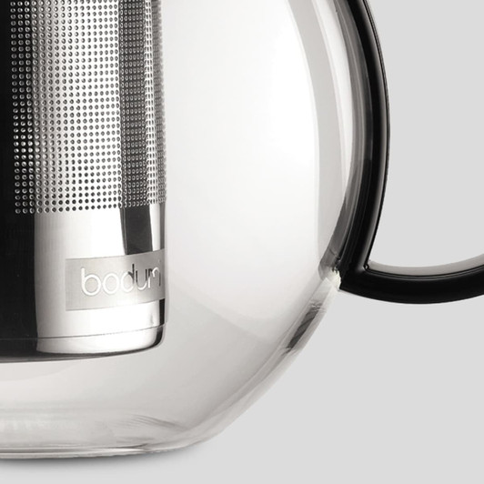 A glass tea press featuring a stainless steel infuser and a black handle, with a logo on the body.