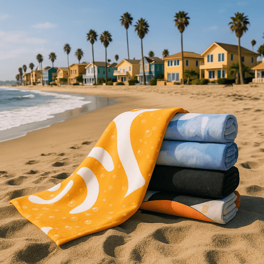 A stack of five bath towels in various colours, including yellow, blue, and black, on a sandy beach with palm trees and houses in the background.