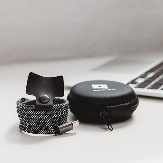 A charging cable in black and white fabric with a snap closure, packaged in a circular black case with a logo.