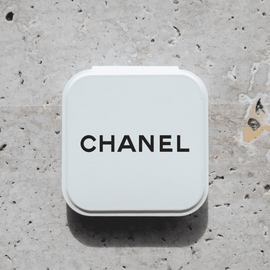 A compact mirror in a white case with a logo on the top, set against a textured stone background.