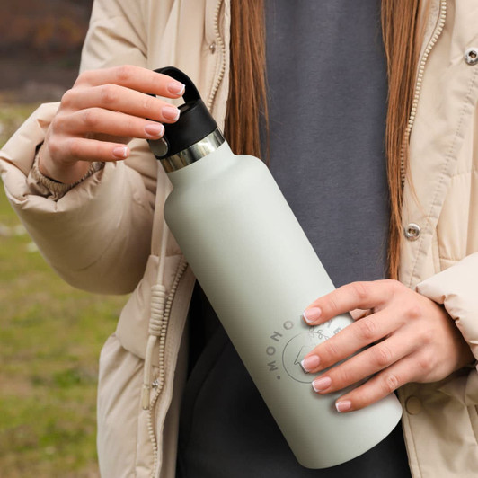 A drink bottle in a soft green colour with a carry lid, held by a person wearing a light jacket in an outdoor setting.