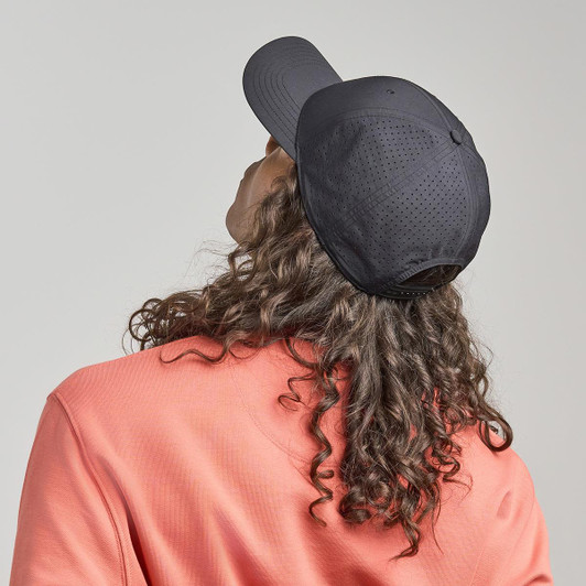 A black cap is worn backwards on the head of a person with curly hair, set against a neutral background.
