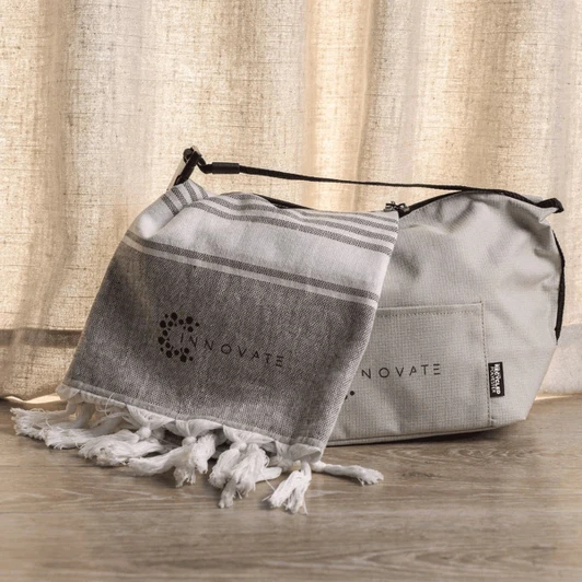 A grey cooler bag with a matching grey and white towel featuring tassels, both displaying branding.