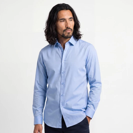 A man in a light blue long-sleeve shirt stands against a neutral background. The shirt features a collar and buttons.