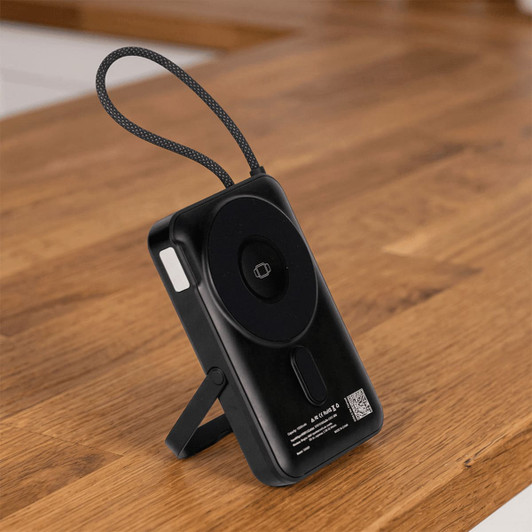 A black power bank with a magnetic feature and a stand, set on a wooden surface. It includes a textured strap for easy handling.