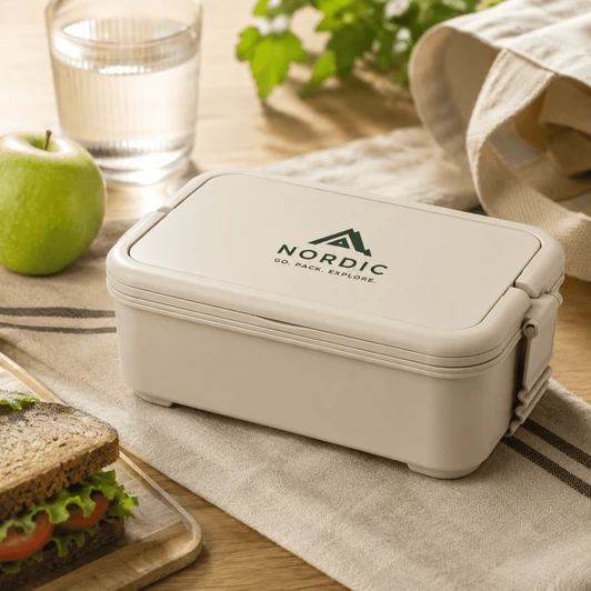 A beige lunchbox with a logo, placed on a table beside a sandwich, an apple, a glass of water, and a cloth.