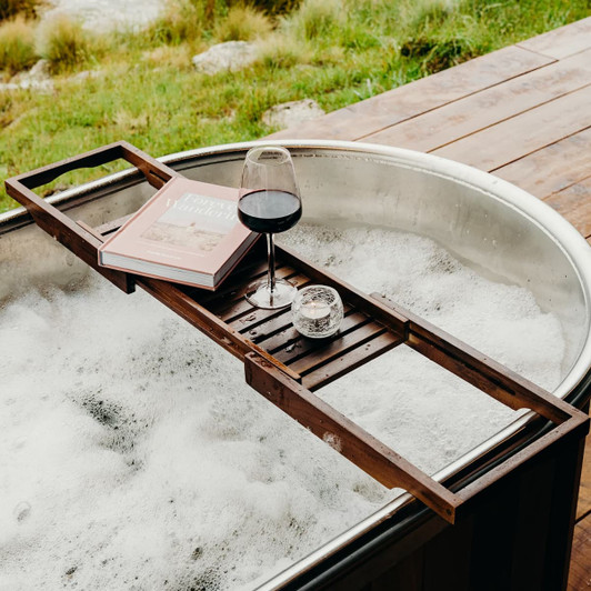 A wooden bath caddy in dark brown, holding a glass of red wine, a candle, and a book, resting over a bubbly bathtub.