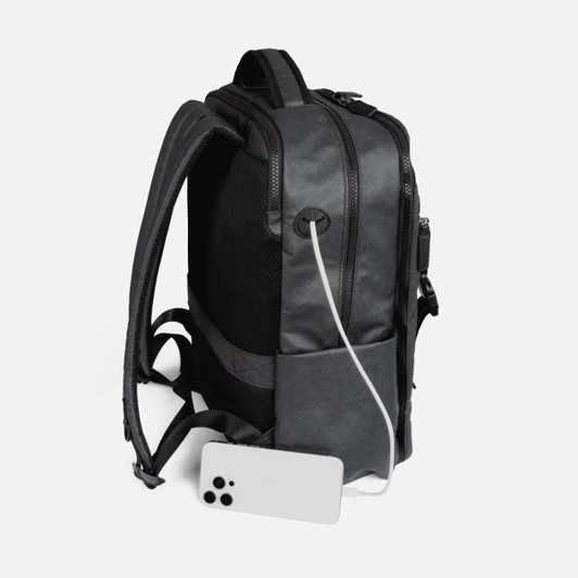 A black utility backpack with a charging cable and a smartphone at its base. It features a logo on the side.