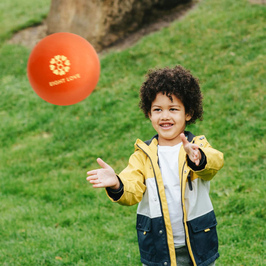 An inflatable PVC ball in orange, held by a smiling child wearing a yellow and navy jacket, on green grass.