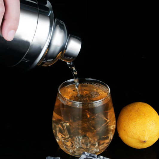 A stainless steel cocktail shaker pouring a drink into a glass with ice, accompanied by a lemon on a black background.