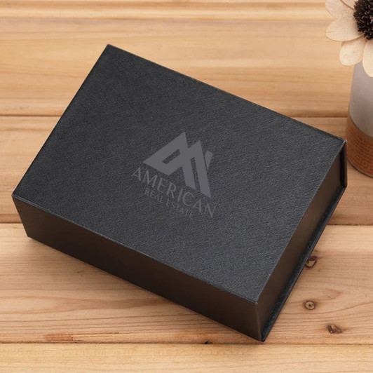 A black magnetic gift box with a textured finish and a logo on the top, placed on a wooden surface.