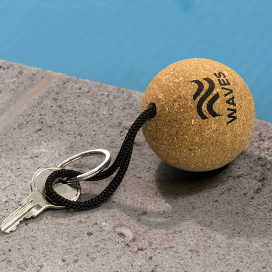 A round cork key ring with a black cord and metal keyring, featuring a logo. Set against a textured stone surface.