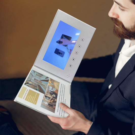 A white 7-inch LCD video brochure with an open cover displaying a video on the screen, held by a man in formal attire.