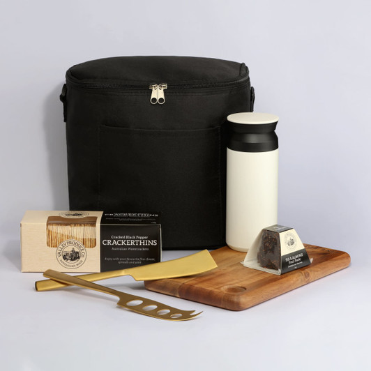A black cooler bag with a drink bottle, wooden serving board, cheese knife, and packaged cracker thins.