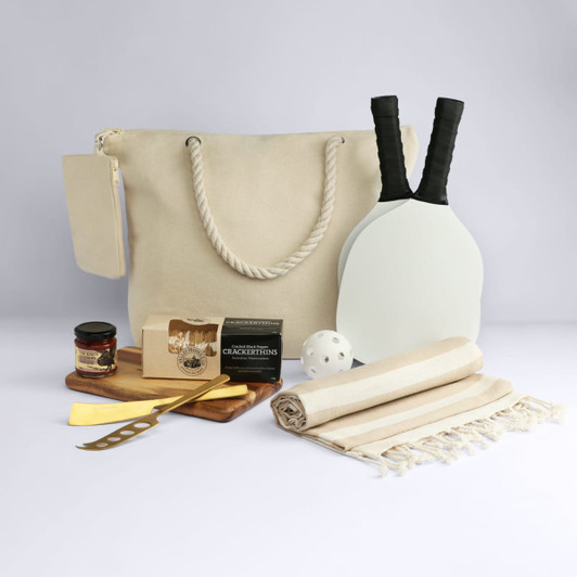 A beige tote bag with rope handles, picnic utensils, a paddle set, a ball, a cheese board, and a jar of jam.