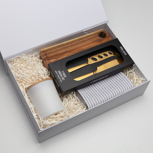 A gift box containing a wooden cheese board, gold knife, a glass cup, and a grey striped towel.