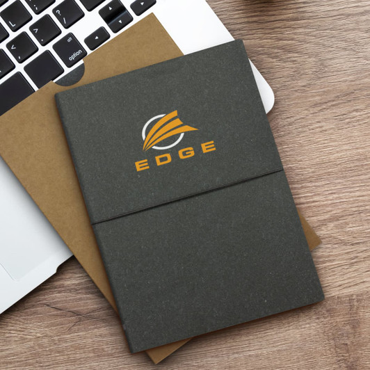 A grey recycled soft cover notebook with an orange logo, placed on a wooden surface next to a laptop.