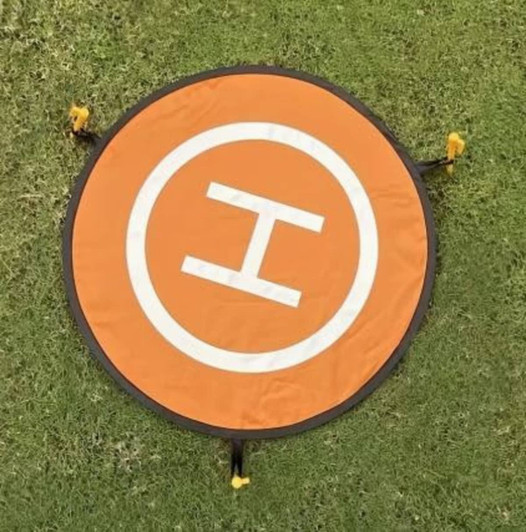An orange drone landing mat with a white "H" symbol, secured to the ground with yellow ties.