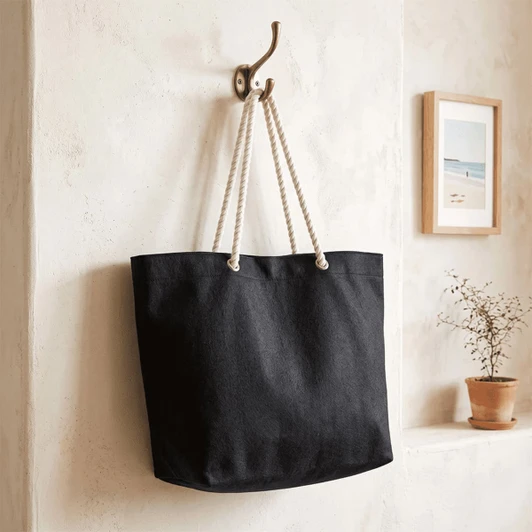 A black recycled beach tote bag with rope handles hangs on a hook against a textured wall.