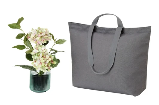A grey tote bag with two long handles beside a vase with decorative flowers. The bag features a logo.